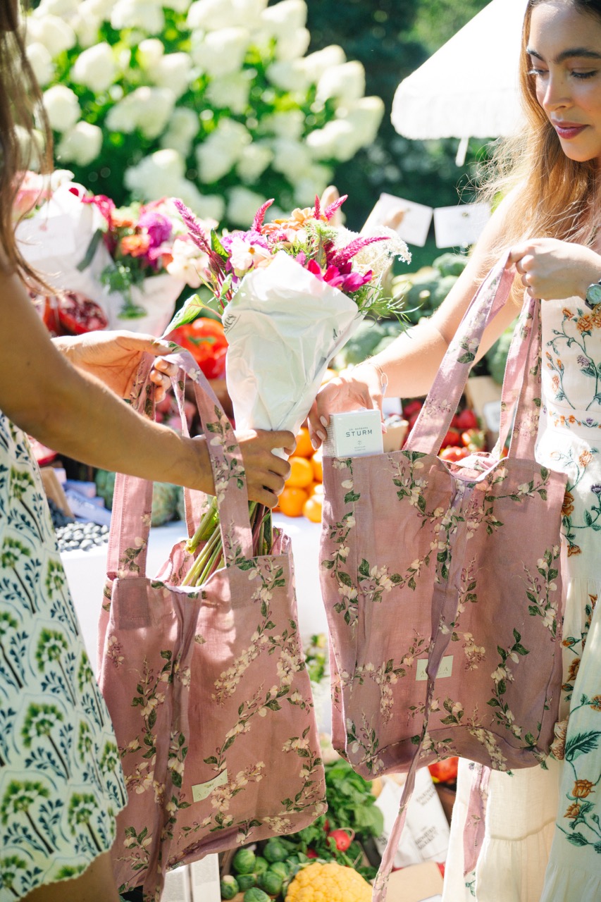 Guests fill floral tote bags with fresh flowers and curated gifts at the Agua by Agua Bendita x CAKE farmer's market brand activation in the Hamptons, surrounded by colorful produce and floral displays.